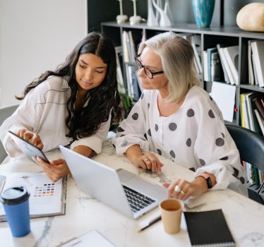 Two professional women discussing work on a laptop and tablet in a contemporary office setting.